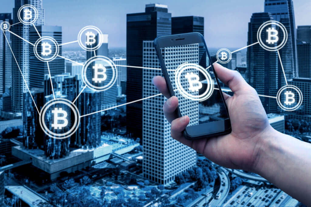 Celular muestra el símbolo de bitcoin y de fondo edificios y propiedades inmobiliarias.